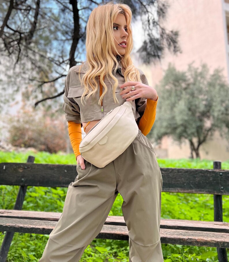 May include: A woman wearing a khaki green cropped jacket and matching pants with a white fanny pack. She is standing in front of a wooden bench with green grass behind her.