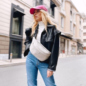 May include: A woman wearing a black leather jacket with a white turtleneck sweater, blue jeans, and a pink baseball cap. She is carrying a light gray fanny pack.