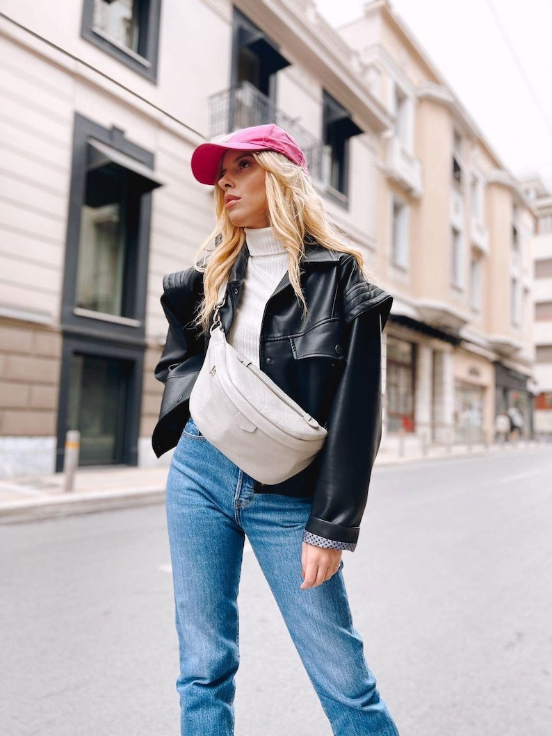 May include: A woman wearing a black leather jacket with a white turtleneck sweater, blue jeans, and a pink baseball cap. She is carrying a light gray fanny pack.