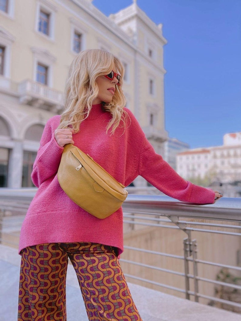 May include: A woman wearing a pink sweater and patterned pants with a mustard yellow fanny pack.