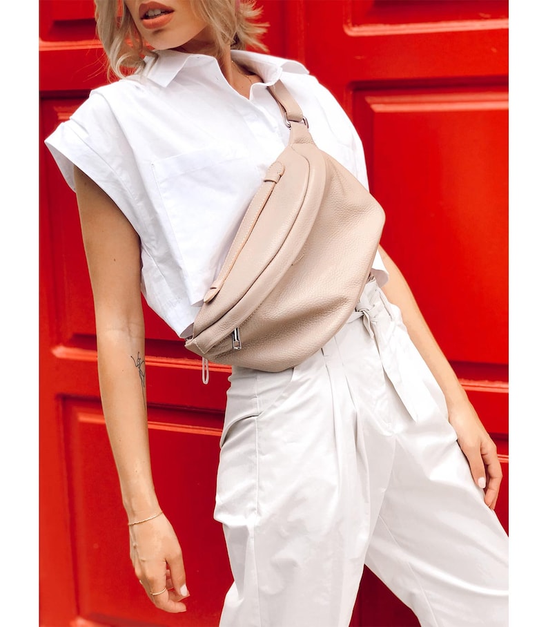 May include: A light beige leather fanny pack with a silver zipper and buckle. The fanny pack is worn around the waist of a person wearing a white shirt and white pants.