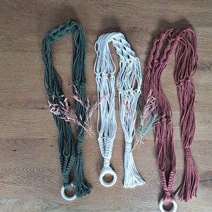 Könnte beinhalten: Drei Makramee-Pflanzenhänger in verschiedenen Farben: grün, weiß und rosa. Jeder Hänger hat einen Holzring oben und einen Quasten unten.