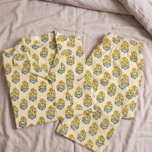 K&ouml;nnte beinhalten: Cremefarbenes Pyjama-Set mit einem gelb-blauen Blumenmuster. Das Set beinhaltet ein Hemd mit Kn&ouml;pfen und eine passende Hose. Die Pyjamas sind aus einem weichen Stoff und liegen auf einem Bett.