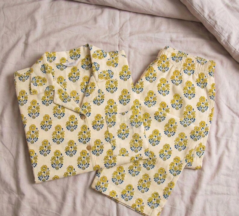 K&ouml;nnte beinhalten: Cremefarbenes Pyjama-Set mit einem gelb-blauen Blumenmuster. Das Set beinhaltet ein Hemd mit Kn&ouml;pfen und eine passende Hose. Die Pyjamas sind aus einem weichen Stoff und liegen auf einem Bett.