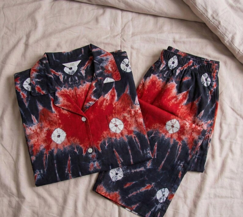K&ouml;nnte beinhalten: Ein Schlafanzug-Set im Batik-Design in Rot, Marineblau und Wei&szlig;. Das Set beinhaltet ein Hemd mit Kn&ouml;pfen und eine passende Hose. Das Hemd hat einen Kragen und die Hose hat einen elastischen Bund. Der Stoff hat ein einzigartiges, k&uuml;nstlerisches Muster.