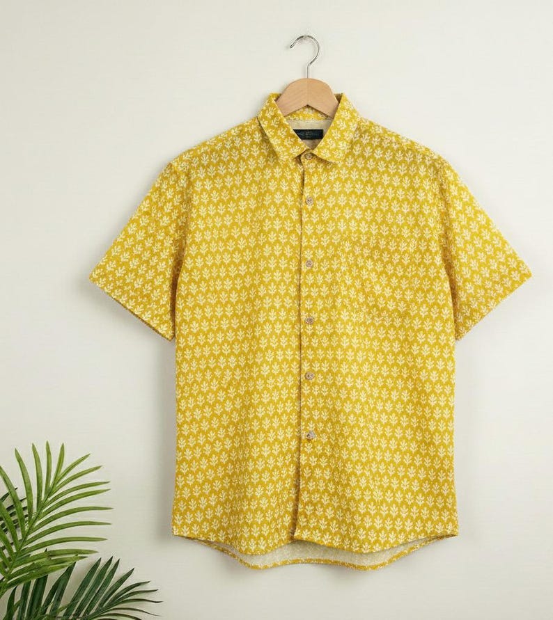 Puede incluir: Camisa amarilla mostaza de manga corta con un estampado de hojas blancas. La camisa tiene una botonadura, un cuello y un bolsillo en el pecho. Cuelga de una percha de madera sobre un fondo blanco.