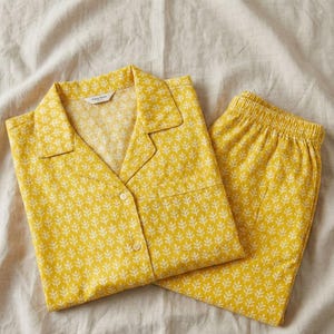 K&ouml;nnte beinhalten: Gelbes Pyjama-Set mit wei&szlig;em Bl&auml;ttermuster. Das Set besteht aus einem Hemd mit Kn&ouml;pfen und Brusttasche sowie einer passenden Hose mit elastischem Bund. Der Pyjama ist aus weichem Stoff gefertigt und liegt gefaltet auf einem neutralfarbenen Laken.