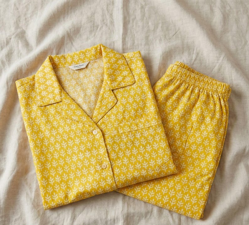 K&ouml;nnte beinhalten: Gelbes Pyjama-Set mit wei&szlig;em Bl&auml;ttermuster. Das Set besteht aus einem Hemd mit Kn&ouml;pfen und Brusttasche sowie einer passenden Hose mit elastischem Bund. Der Pyjama ist aus weichem Stoff gefertigt und liegt gefaltet auf einem neutralfarbenen Laken.