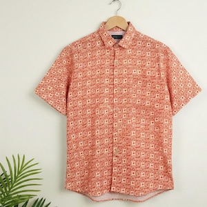 Puede incluir: Una camisa de manga corta de color coral con un patr&oacute;n geom&eacute;trico crema repetido. La camisa est&aacute; colgada de una percha de madera. Tiene cuello y bolsillo. La tela es ligera e ideal para el verano.