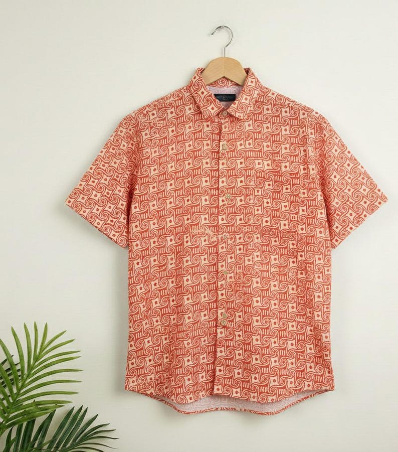 Puede incluir: Una camisa de manga corta de color coral con un patr&oacute;n geom&eacute;trico crema repetido. La camisa est&aacute; colgada de una percha de madera. Tiene cuello y bolsillo. La tela es ligera e ideal para el verano.