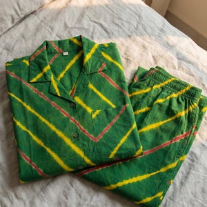 K&ouml;nnte beinhalten: Gr&uuml;nes Pyjama-Set mit gelben und roten diagonalen Streifen. Das Oberteil hat einen Kragen, eine Vordertasche und Kn&ouml;pfe. Die passende Hose hat einen elastischen Bund. Der Stoff scheint Baumwolle zu sein.