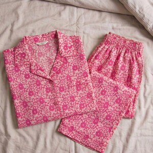 K&ouml;nnte beinhalten: Rosa Pyjama-Set mit Blumenmuster. Das Set besteht aus einem Hemd mit Kn&ouml;pfen und Kragen sowie einer passenden Hose. Die Pyjamas sind aus einem weichen Stoff gefertigt und weisen ein sich wiederholendes Blumenmuster in Rosa- und Wei&szlig;t&ouml;nen auf. Das Set liegt auf einem wei&szlig;en Laken.