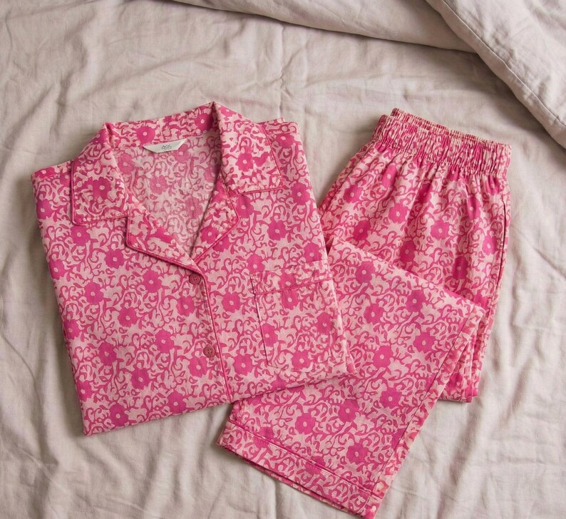 K&ouml;nnte beinhalten: Rosa Pyjama-Set mit Blumenmuster. Das Set besteht aus einem Hemd mit Kn&ouml;pfen und Kragen sowie einer passenden Hose. Die Pyjamas sind aus einem weichen Stoff gefertigt und weisen ein sich wiederholendes Blumenmuster in Rosa- und Wei&szlig;t&ouml;nen auf. Das Set liegt auf einem wei&szlig;en Laken.