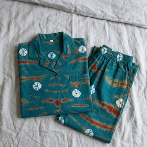 K&ouml;nnte beinhalten: T&uuml;rkis- und rostfarbenes Pyjama-Set mit einem Hemd mit Kn&ouml;pfen und passender Hose. Das Set hat ein Batikmuster mit wei&szlig;en kreisf&ouml;rmigen Designs. Das Hemd hat eine Brusttasche. Das Pyjama-Set wird auf einem grauen Bettlaken pr&auml;sentiert.
