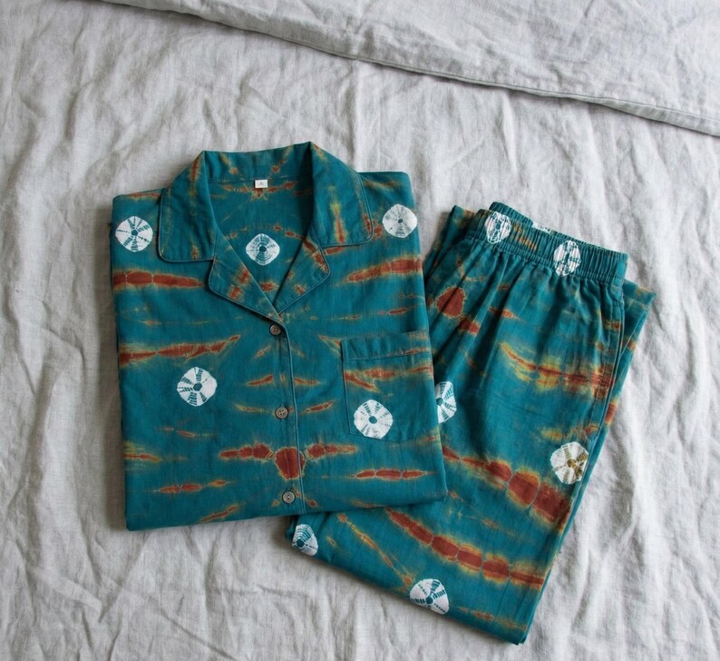 K&ouml;nnte beinhalten: T&uuml;rkis- und rostfarbenes Pyjama-Set mit einem Hemd mit Kn&ouml;pfen und passender Hose. Das Set hat ein Batikmuster mit wei&szlig;en kreisf&ouml;rmigen Designs. Das Hemd hat eine Brusttasche. Das Pyjama-Set wird auf einem grauen Bettlaken pr&auml;sentiert.