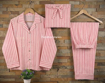 Könnte beinhalten: Ein rosa-weiß gestreiftes Pyjama-Set, bestehend aus einem Hemd mit Knöpfen, einer Hose und einem passenden Beutel. Das Hemd hat eine Brusttasche und lange Ärmel. Die Hose hat einen elastischen Bund. Das Set wird auf einem Holzbügel präsentiert.