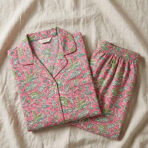 K&ouml;nnte beinhalten: Rosa Pyjama-Set mit floralem Muster in Gr&uuml;n und Wei&szlig;. Das Set besteht aus einem Oberteil mit Kn&ouml;pfen und Reverskragen sowie einer passenden Hose mit elastischem Bund. Der Stoff wirkt weich und leicht, geeignet f&uuml;r Nachtw&auml;sche.