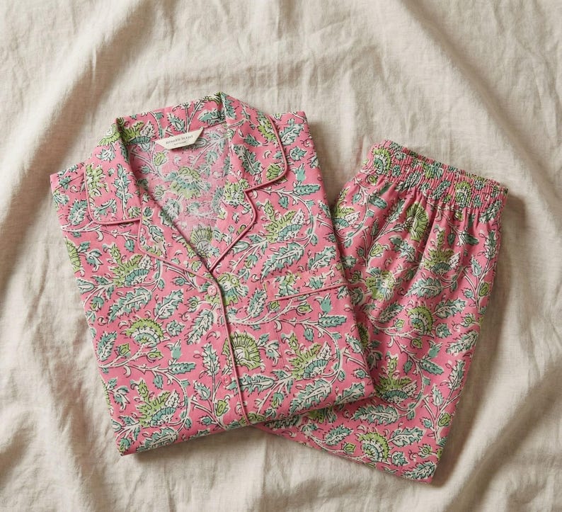 K&ouml;nnte beinhalten: Rosa Pyjama-Set mit floralem Muster in Gr&uuml;n und Wei&szlig;. Das Set besteht aus einem Oberteil mit Kn&ouml;pfen und Reverskragen sowie einer passenden Hose mit elastischem Bund. Der Stoff wirkt weich und leicht, geeignet f&uuml;r Nachtw&auml;sche.