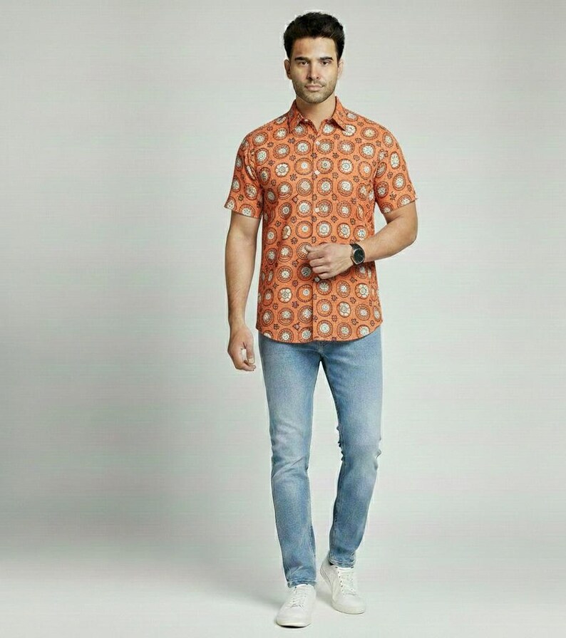 Puede incluir: Una camisa naranja de manga corta con un patr&oacute;n circular repetido en blanco y marr&oacute;n, combinada con vaqueros azul claro y zapatillas blancas. La camisa tiene un cuello cl&aacute;sico y se lleva con un reloj negro.