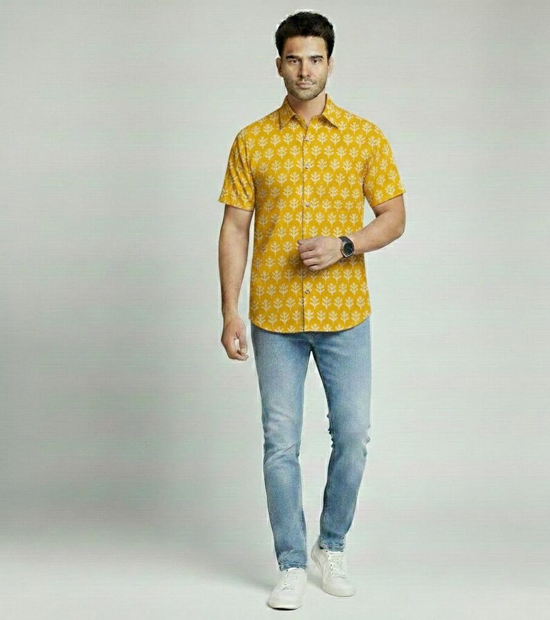 Puede incluir: Camisa amarilla mostaza de manga corta con un estampado de hojas blancas. La camisa se combina con vaqueros azul claro y zapatillas blancas. Un reloj se lleva en la mu&ntilde;eca izquierda. El conjunto es informal.