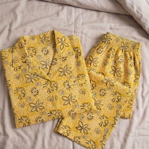 K&ouml;nnte beinhalten: Gelbes Pyjama-Set mit floralem Muster. Das Set beinhaltet ein Hemd mit Kn&ouml;pfen und Reverskragen sowie eine Hose mit elastischem Bund. Das florale Design weist dunkelbraune und wei&szlig;e Akzente auf gelbem Hintergrund auf.
