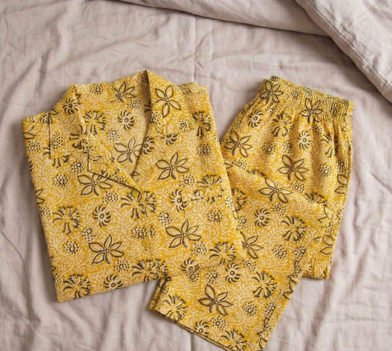 K&ouml;nnte beinhalten: Gelbes Pyjama-Set mit floralem Muster. Das Set beinhaltet ein Hemd mit Kn&ouml;pfen und Reverskragen sowie eine Hose mit elastischem Bund. Das florale Design weist dunkelbraune und wei&szlig;e Akzente auf gelbem Hintergrund auf.