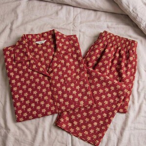 K&ouml;nnte beinhalten: Rotes Pyjama-Set mit sich wiederholendem goldenem Blumenmuster. Das Set beinhaltet ein Hemd mit Kn&ouml;pfen und einer Brusttasche sowie eine passende Hose. Der Pyjama ist aus weichem Stoff und liegt auf einem Bett.