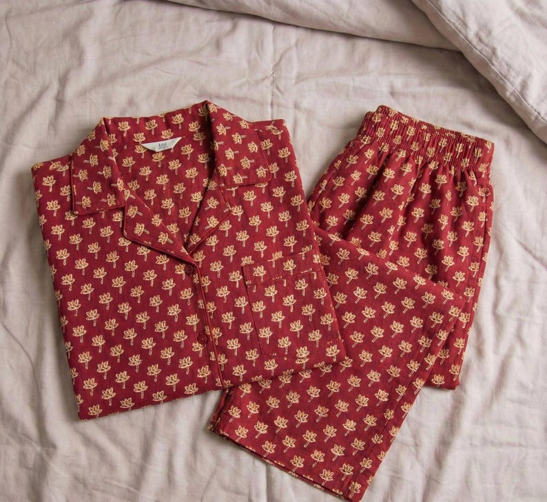 K&ouml;nnte beinhalten: Rotes Pyjama-Set mit sich wiederholendem goldenem Blumenmuster. Das Set beinhaltet ein Hemd mit Kn&ouml;pfen und einer Brusttasche sowie eine passende Hose. Der Pyjama ist aus weichem Stoff und liegt auf einem Bett.
