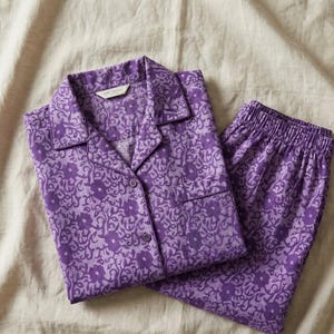 K&ouml;nnte beinhalten: Lila Pyjama-Set mit floralem Muster. Das Set besteht aus einem Hemd mit Kn&ouml;pfen und einer Brusttasche sowie einer passenden Hose mit elastischem Bund. Der Stoff hat ein helllila Blumenmuster auf einem dunkleren lilafarbenen Hintergrund.