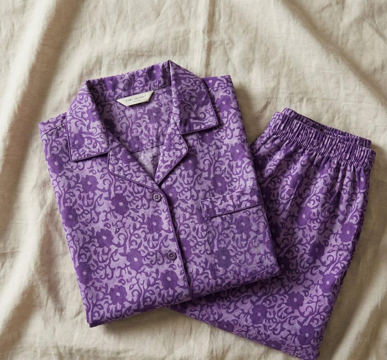 K&ouml;nnte beinhalten: Lila Pyjama-Set mit floralem Muster. Das Set besteht aus einem Hemd mit Kn&ouml;pfen und einer Brusttasche sowie einer passenden Hose mit elastischem Bund. Der Stoff hat ein helllila Blumenmuster auf einem dunkleren lilafarbenen Hintergrund.