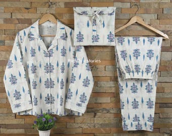 Könnte beinhalten: Ein weißes Pyjama-Set mit blauem Blumen- und Baummuster. Das Set umfasst ein Hemd mit Knöpfen, eine Hose und eine passende Kordelzugtasche. Die Pyjamas werden auf einem Holzbügel vor einer Backsteinmauer präsentiert.