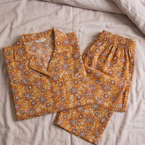 K&ouml;nnte beinhalten: Ein zweiteiliger Schlafanzug mit floralem Muster in Senfgelb. Das Set besteht aus einem Hemd mit Kn&ouml;pfen und einer Hose. Das Muster zeigt wei&szlig;e und bordeauxfarbene Blumen. Der Schlafanzug liegt auf einem Bett.