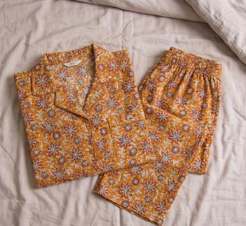 K&ouml;nnte beinhalten: Ein zweiteiliger Schlafanzug mit floralem Muster in Senfgelb. Das Set besteht aus einem Hemd mit Kn&ouml;pfen und einer Hose. Das Muster zeigt wei&szlig;e und bordeauxfarbene Blumen. Der Schlafanzug liegt auf einem Bett.