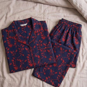 K&ouml;nnte beinhalten: Marineblaues Pyjama-Set mit rotem Blumenmuster. Das Set besteht aus einem Hemd mit Kn&ouml;pfen und einem Reverskragen sowie einer passenden Hose. Die Kanten des Oberteils und der Tasche sind rot eingefasst.