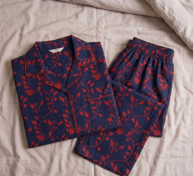 K&ouml;nnte beinhalten: Marineblaues Pyjama-Set mit rotem Blumenmuster. Das Set besteht aus einem Hemd mit Kn&ouml;pfen und einem Reverskragen sowie einer passenden Hose. Die Kanten des Oberteils und der Tasche sind rot eingefasst.