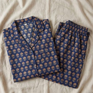 K&ouml;nnte beinhalten: Marineblaues Pyjama-Set mit einem sich wiederholenden Blumenmuster in Goldbraun. Das Set besteht aus einem Oberteil mit Kn&ouml;pfen und Kragen sowie einer passenden Hose. Der Stoff wirkt weich und leicht, geeignet f&uuml;r Nachtw&auml;sche.