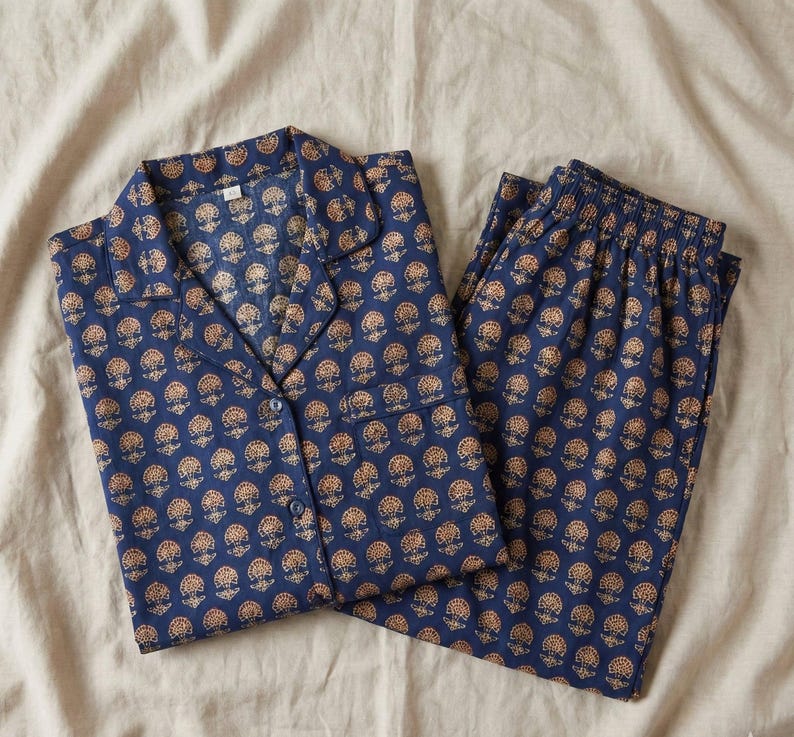 K&ouml;nnte beinhalten: Marineblaues Pyjama-Set mit einem sich wiederholenden Blumenmuster in Goldbraun. Das Set besteht aus einem Oberteil mit Kn&ouml;pfen und Kragen sowie einer passenden Hose. Der Stoff wirkt weich und leicht, geeignet f&uuml;r Nachtw&auml;sche.