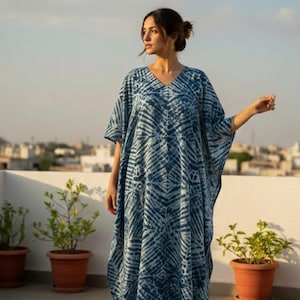 May include: A long, flowing blue and white patterned kaftan dress with a V-neck. The dress has short sleeves and a loose fit. The model is standing on a rooftop with potted plants in the background.