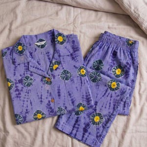 K&ouml;nnte beinhalten: Ein lavendelfarbenes Pyjama-Set mit Batikmuster, bestehend aus einem Hemd mit Kn&ouml;pfen und einer passenden Hose. Das Set ist mit einem sich wiederholenden Muster aus marineblauen und gelben Kreismotiven verziert. Die Pyjamas sind aus einem weichen Stoff gefertigt.