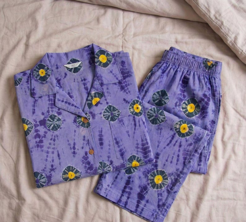 K&ouml;nnte beinhalten: Ein lavendelfarbenes Pyjama-Set mit Batikmuster, bestehend aus einem Hemd mit Kn&ouml;pfen und einer passenden Hose. Das Set ist mit einem sich wiederholenden Muster aus marineblauen und gelben Kreismotiven verziert. Die Pyjamas sind aus einem weichen Stoff gefertigt.
