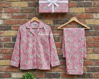 Könnte beinhalten: Rosa Paisley-Pyjama-Set mit passender Geschenkbox, vor einer Backsteinmauer. Der Pyjama besteht aus einem Hemd mit Knöpfen und einer Hose, beide mit weißem Besatz. Die Geschenkbox ist mit passendem Paisley-Papier verpackt und mit einem weißen Band gebunden.