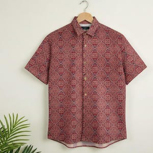 Puede incluir: Una camisa de manga corta con un estampado geom&eacute;trico en tonos rojos, burdeos y azul. La camisa est&aacute; colgada de una percha de madera. El patr&oacute;n es un dise&ntilde;o repetitivo sobre un fondo blanco.