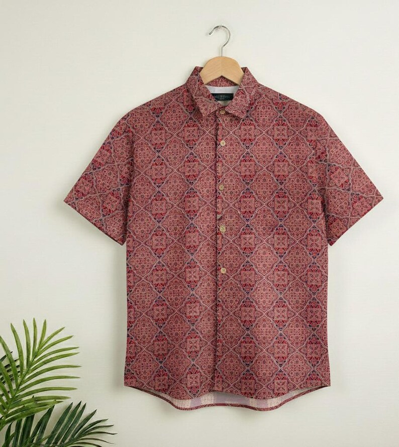 Puede incluir: Una camisa de manga corta con un estampado geom&eacute;trico en tonos rojos, burdeos y azul. La camisa est&aacute; colgada de una percha de madera. El patr&oacute;n es un dise&ntilde;o repetitivo sobre un fondo blanco.