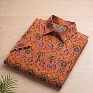 May include: An orange shirt with a floral design in pink and dark grey, neatly folded. The shirt has wooden buttons and a collar with a label that reads "BLOCKPRINT". It is displayed on a light wood surface.