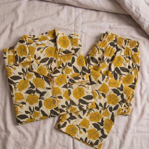 K&ouml;nnte beinhalten: Ein zweiteiliger Schlafanzug in Creme mit einem Blumenmuster. Das Set besteht aus einem Hemd mit Kn&ouml;pfen und Shorts, beide mit gelben Blumen und dunkelgr&uuml;nen Bl&auml;ttern. Der Stoff wirkt weich und bequem.