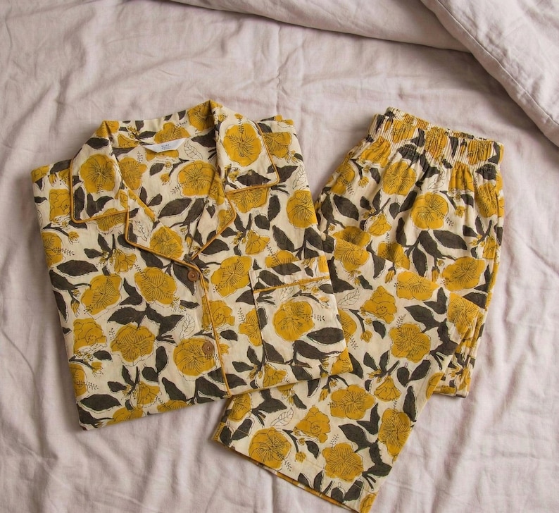 K&ouml;nnte beinhalten: Ein zweiteiliger Schlafanzug in Creme mit einem Blumenmuster. Das Set besteht aus einem Hemd mit Kn&ouml;pfen und Shorts, beide mit gelben Blumen und dunkelgr&uuml;nen Bl&auml;ttern. Der Stoff wirkt weich und bequem.