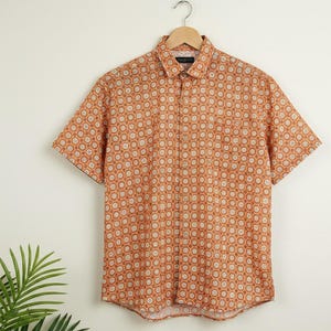 Puede incluir: Camisa de manga corta con un patr&oacute;n geom&eacute;trico repetitivo en naranja y blanco. La camisa est&aacute; colgada de una percha de madera. El cuello y los pu&ntilde;os tienen un patr&oacute;n contrastante. Fondo blanco. Prenda de verano.