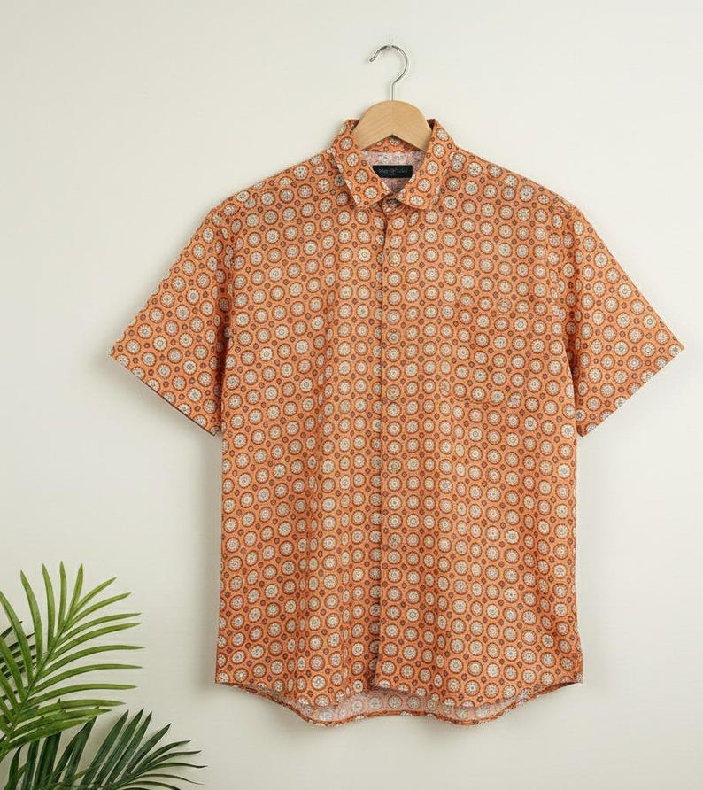 Puede incluir: Camisa de manga corta con un patr&oacute;n geom&eacute;trico repetitivo en naranja y blanco. La camisa est&aacute; colgada de una percha de madera. El cuello y los pu&ntilde;os tienen un patr&oacute;n contrastante. Fondo blanco. Prenda de verano.