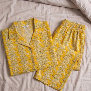 K&ouml;nnte beinhalten: Gelbes Pyjama-Set mit floralem Muster. Das Set besteht aus einem Hemd mit Kn&ouml;pfen und Kragen sowie einer passenden Hose. Der Stoff hat ein wei&szlig;es und gelbes Design. Der Pyjama liegt auf einer hellen Bettdecke.