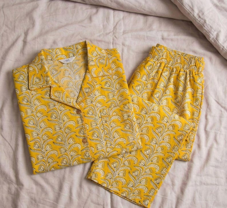 K&ouml;nnte beinhalten: Gelbes Pyjama-Set mit floralem Muster. Das Set besteht aus einem Hemd mit Kn&ouml;pfen und Kragen sowie einer passenden Hose. Der Stoff hat ein wei&szlig;es und gelbes Design. Der Pyjama liegt auf einer hellen Bettdecke.
